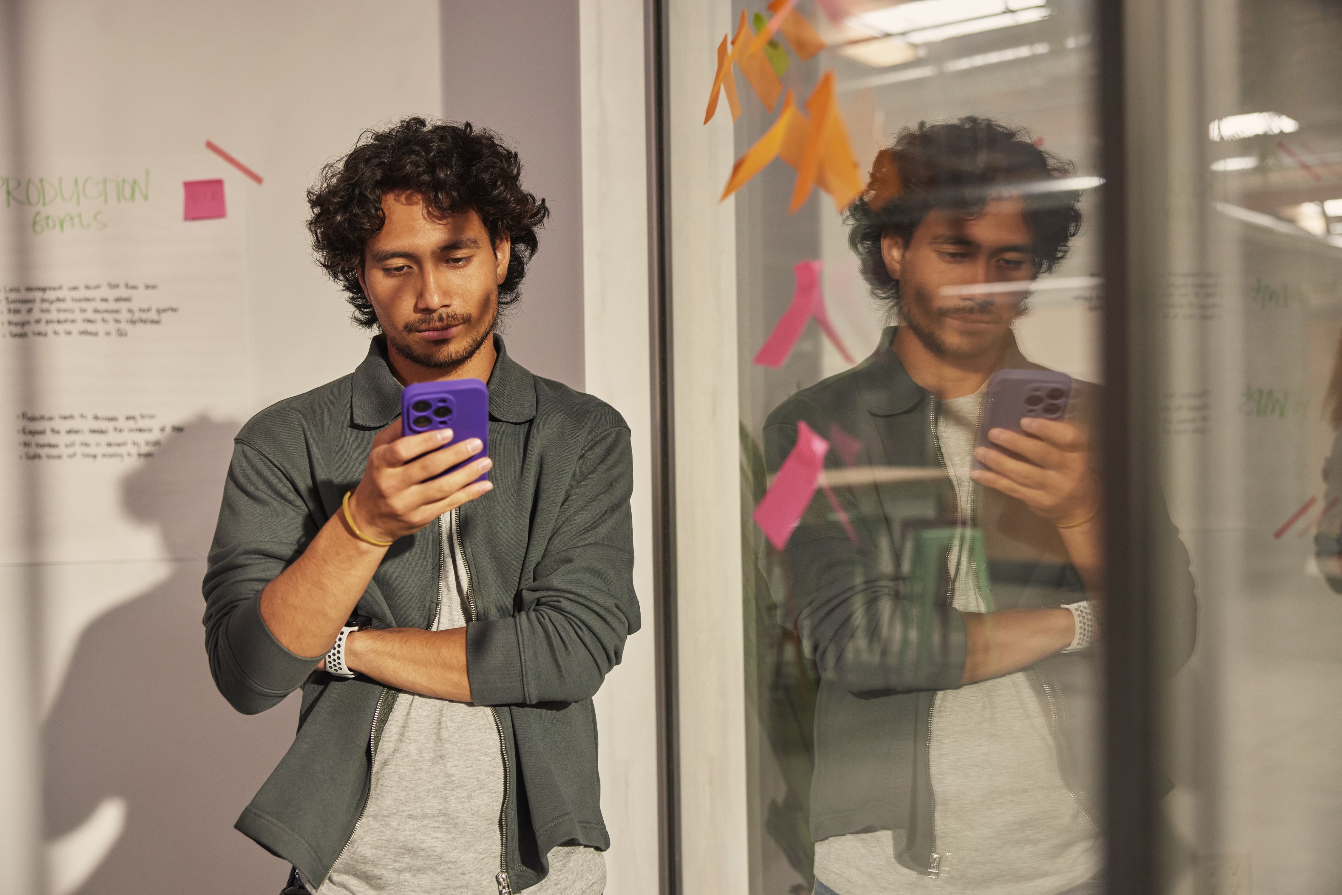 A man photographs himself with a purple smartphone beside a reflective glass wall in a colorful office.