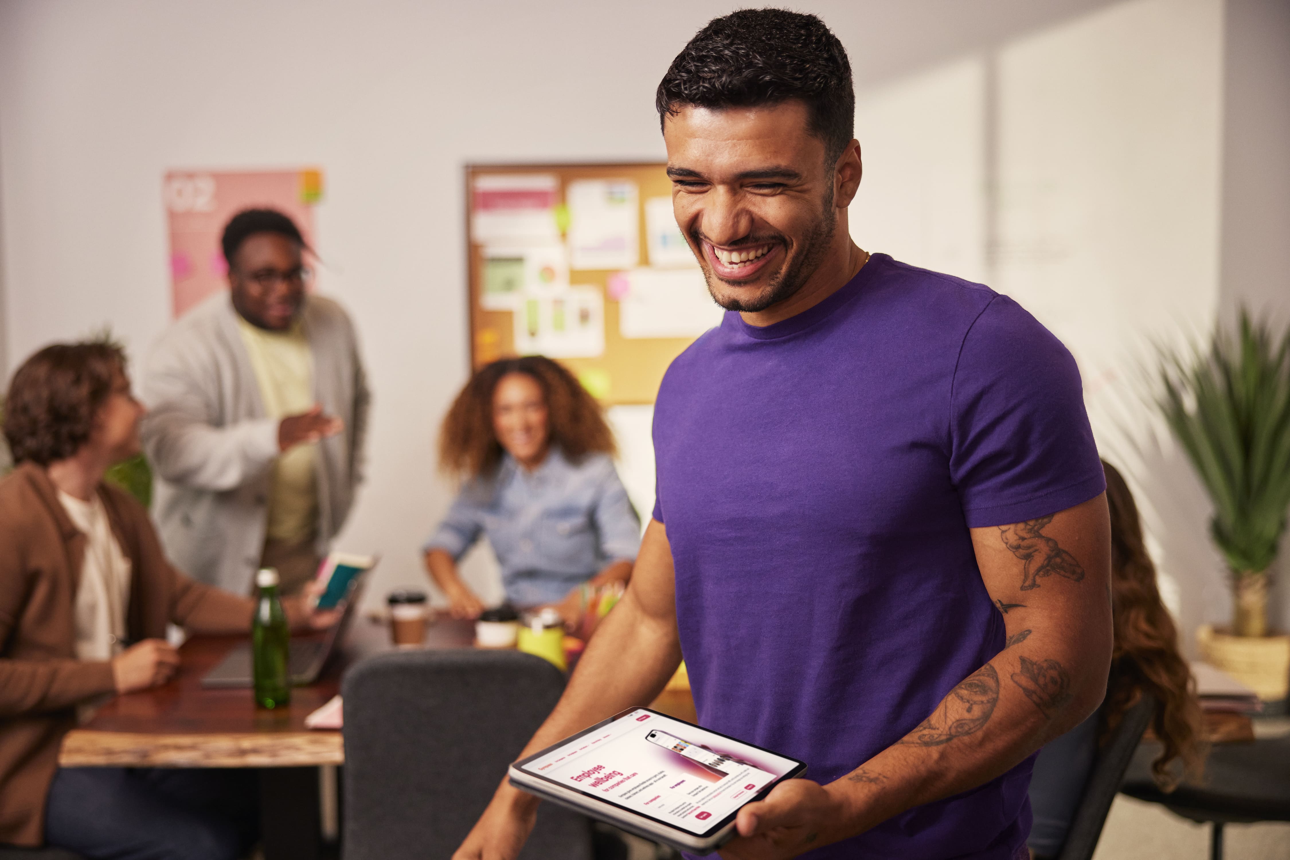 Smiling professional presents a tablet while a diverse team collaborates in a bright creative office.